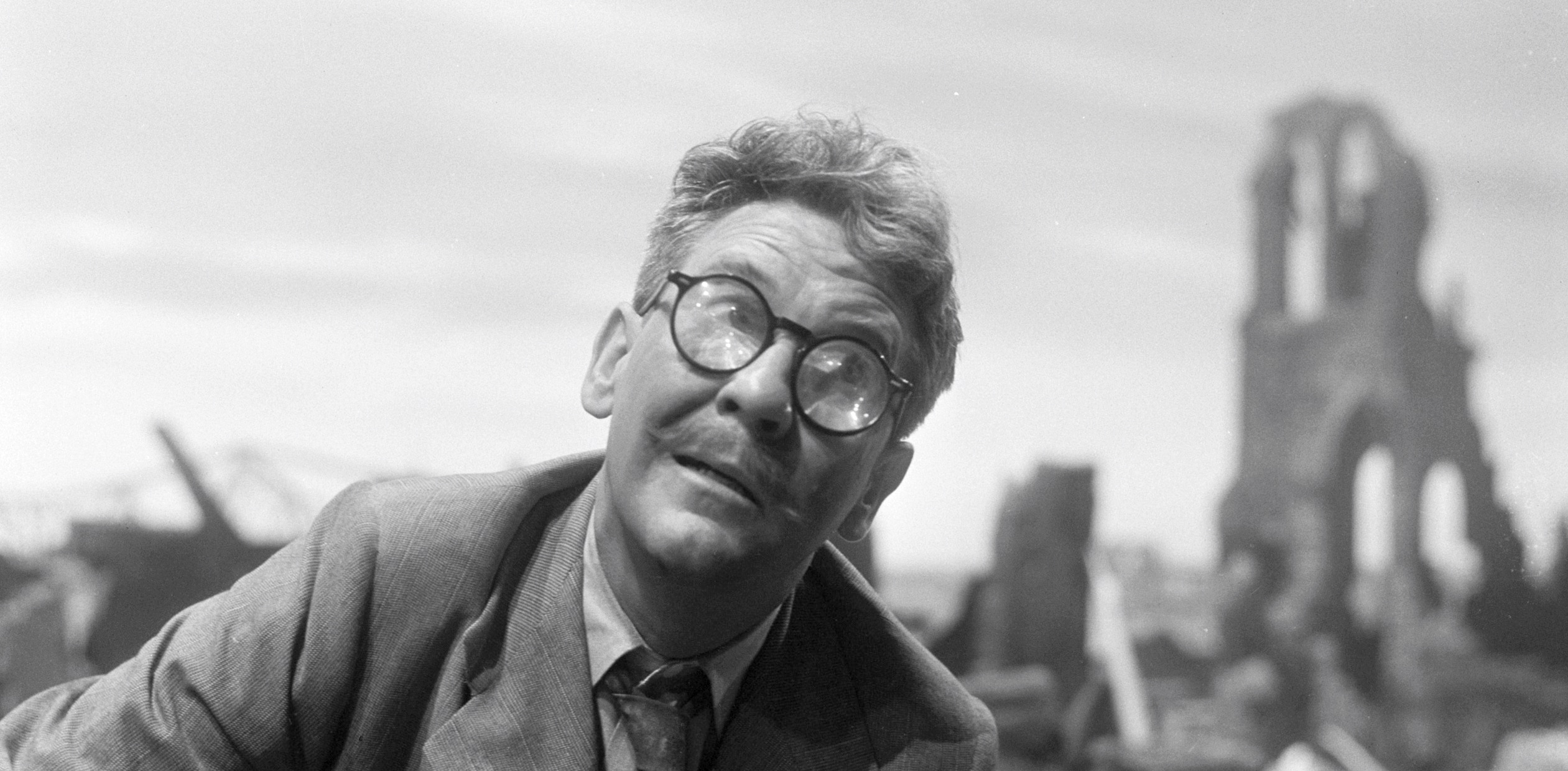 A black and white still photograph from an episode of The Twilight Zone. In the foreground, a man in his 50s leans to one side. His thick glasses are cracked. Behind him, out of focus, is a ruin of some sort.